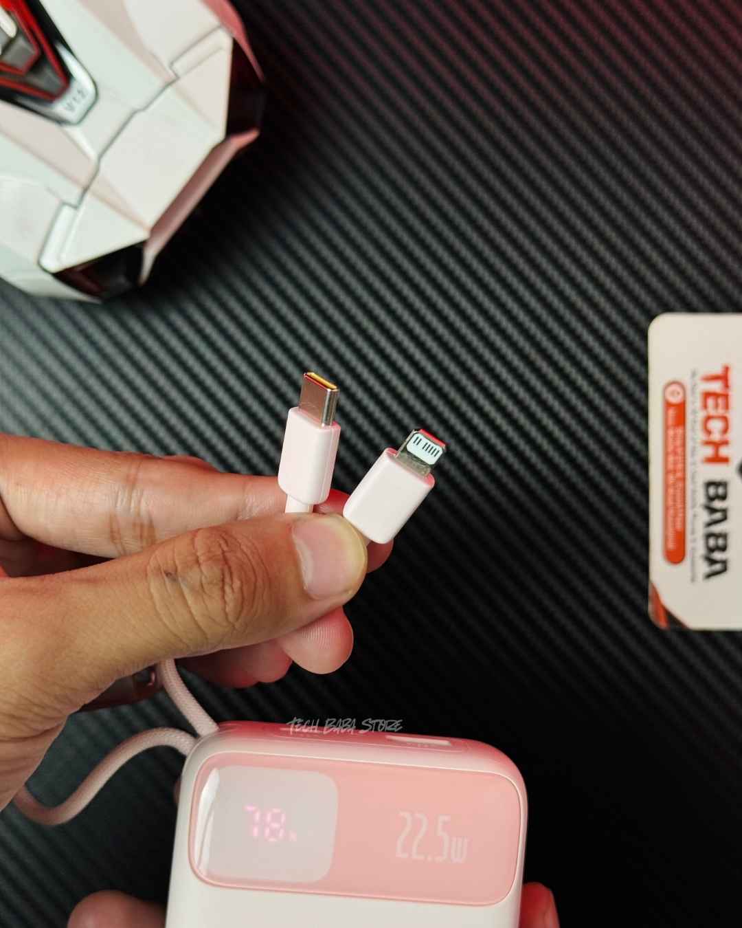 A hand holding a pink colour Baseus Enerfill FC11 20000mAh 22.5W Power Bank showing a closeup of its Built-in 2 Cables which are a Type-C Cable connector and an Lightning port iPhone cable connector, also its showing a front view of powerbank in background which has a digital meter and a label of 22.5W charging speed. Picture by TechBabaStore.pk