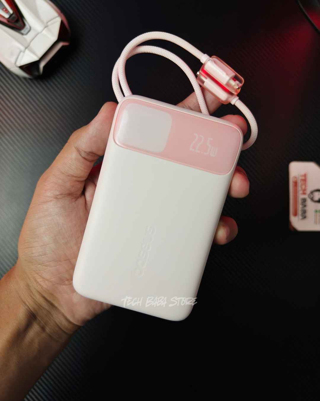 A hand holding a pink colour Baseus Enerfill FC11 20000mAh 22.5W Power Bank showing its 2 built-in cables and a Digital meter located on front and a 22.5W charging speed text label. Picture by TechBabaStore.pk