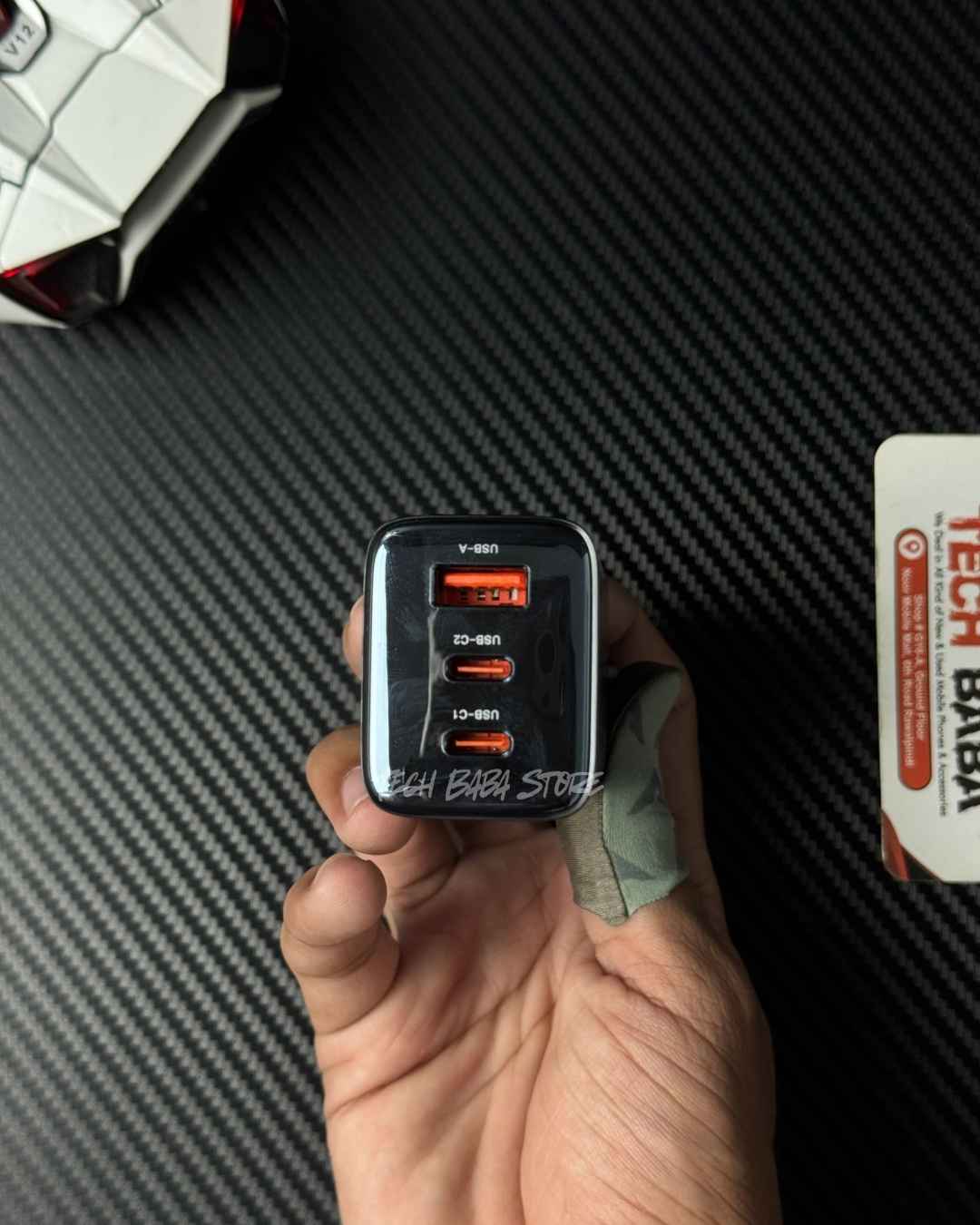 A hand holding a Toocki RX-01 65W 3-port USB power adapter with orange labels, set against a black carbon fiber textured background.