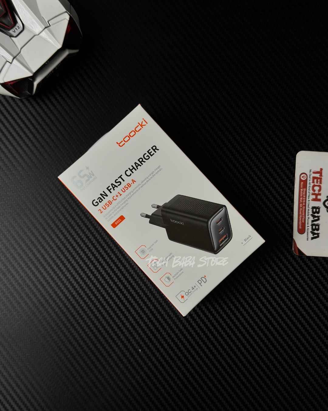 A boxed Toocki RX01 GaN Fast Charger 65W USB-C adapter package placed on a dark textured surface, with part of another product visible at the top left and a small card at the right edge.