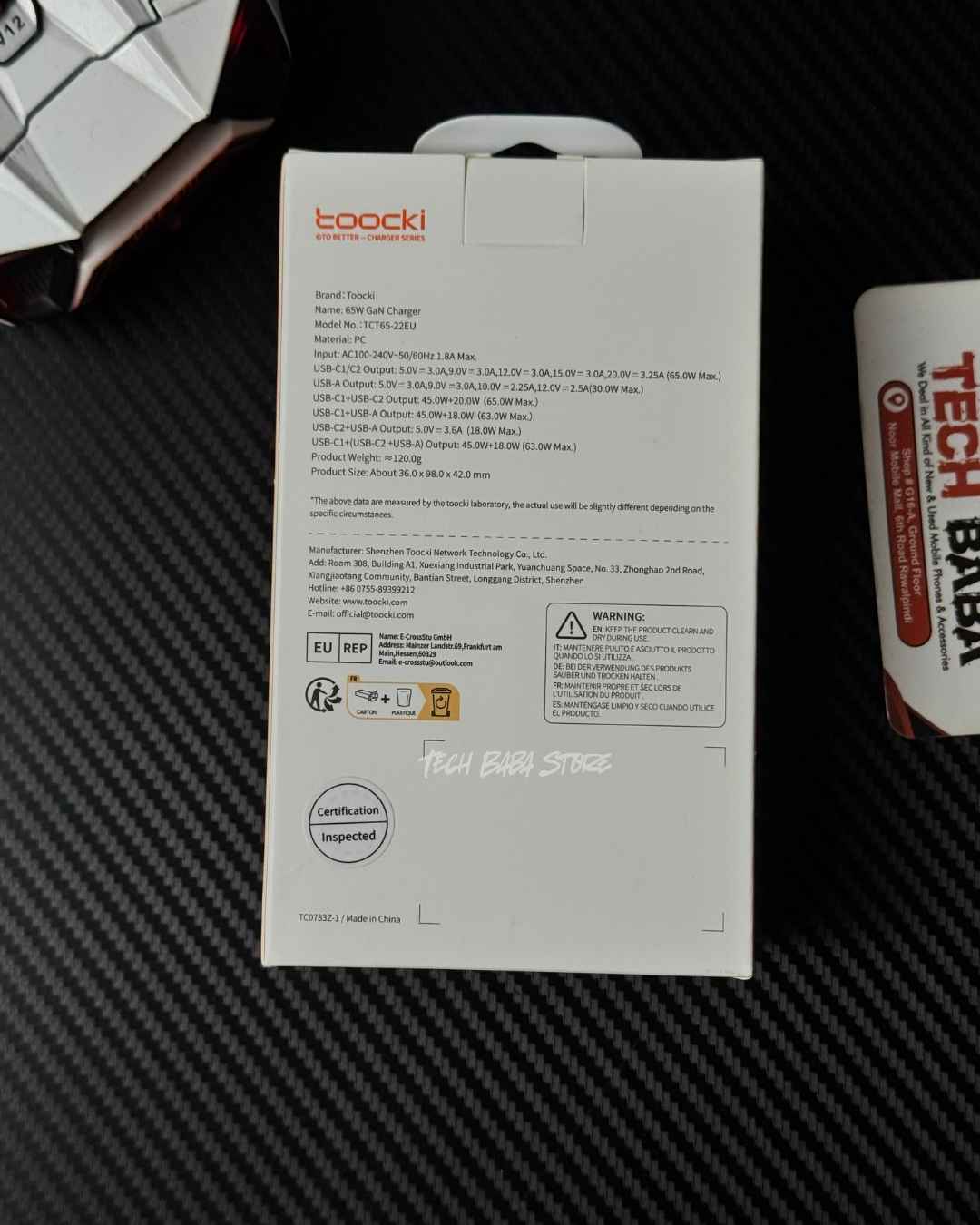 The back of a Toocki 65W RX-01 GaN Adapter product box, showing specifications and text, placed on a black carbon fiber textured surface next to a Tech Breathe tube.