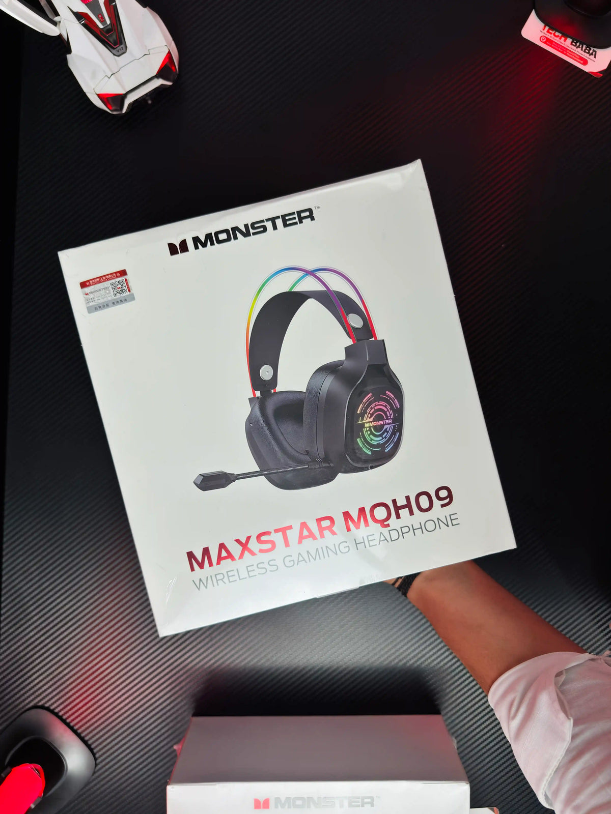 A hand holding black Monster Maxstar MQH09 Wireless Gaming Headphone — 50mm Driver, Bluetooth 5.4, IPX5 | Best PUBG Headset Pakistan which has showing box.pictre by TechBabaStore.pk