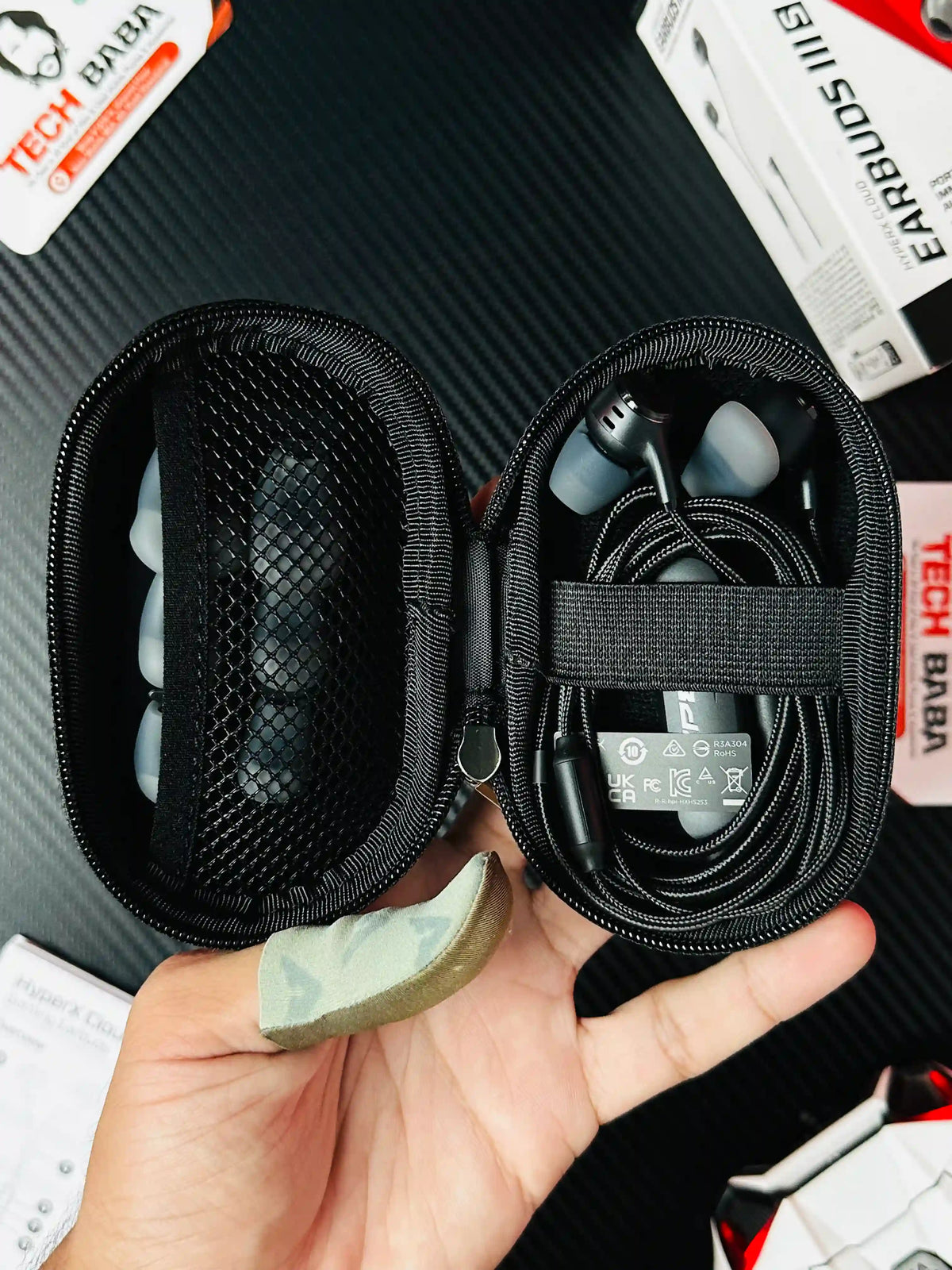 A hand holding open pouch of HyperX Cloud Earbuds III S USB-C Gaming Earbuds which has showing earbuds and buds -picture by TechBabaStore.pk