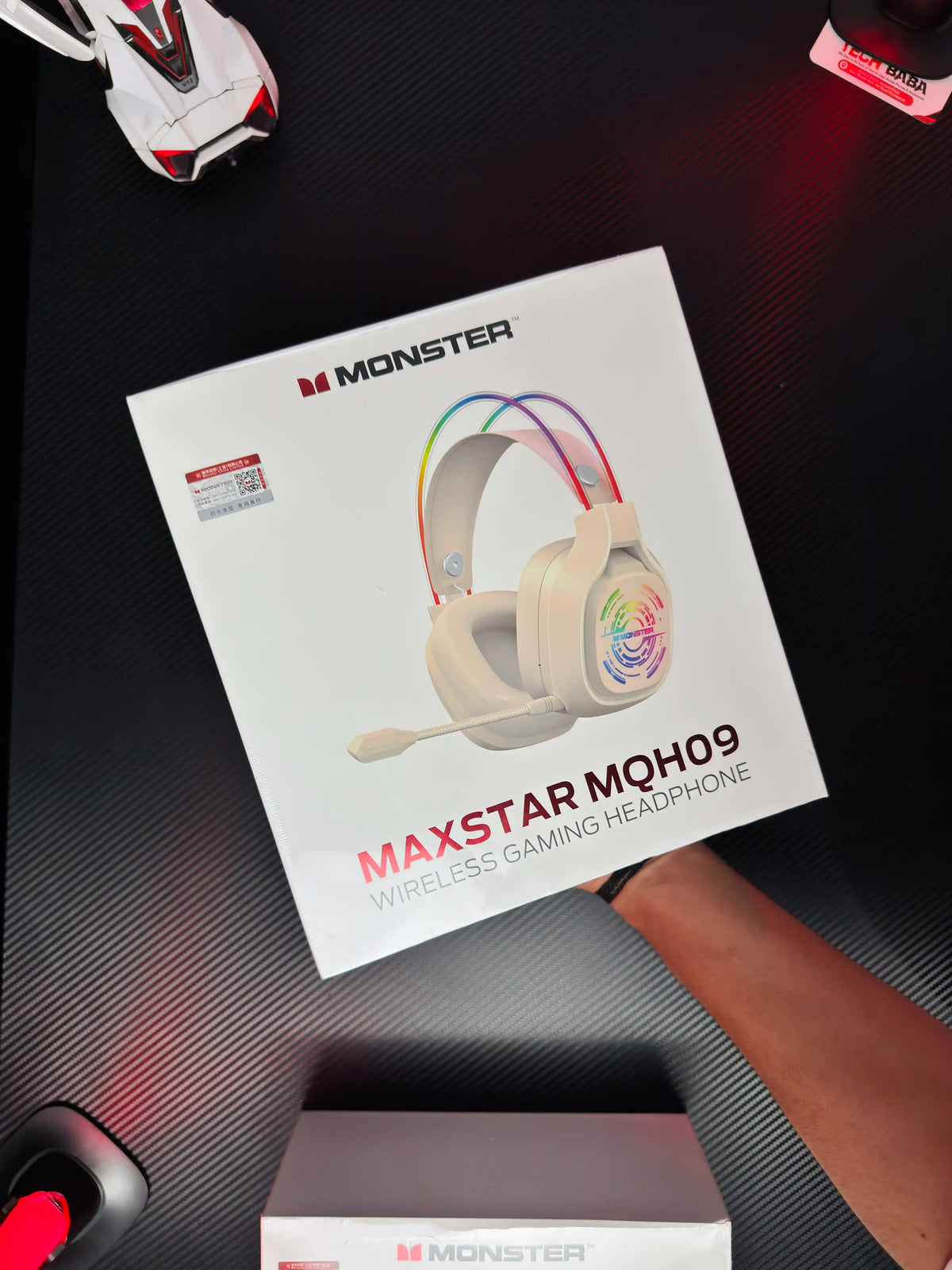 A hand holding white Monster Maxstar MQH09 Wireless Gaming Headphone — 50mm Driver, Bluetooth 5.4, IPX5 | Best PUBG Headset Pakistan which has showing box.pictre by TechBabaStore.pk